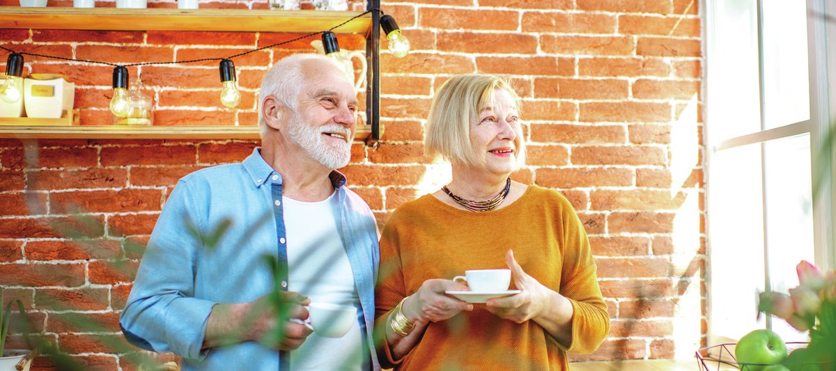 An older couple smiling, holding cups of tea in a bright kitchen